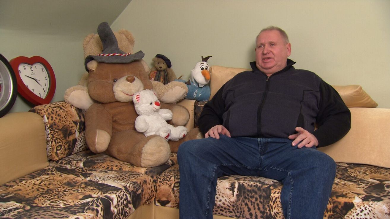 A man sits on a couch with various stuffed animals, including a bear wearing a hat, a white teddy bear, and an Olaf figurine.