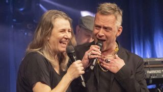 A woman and a man hold microphones and sing together on stage, smiling and enjoying the moment.