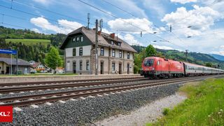 Ein roter Zug fährt durch einen Bahnhof. Einige Leute gehen auf dem Bahnsteig. Im Hintergrund befinden sich Bäume und Berge.