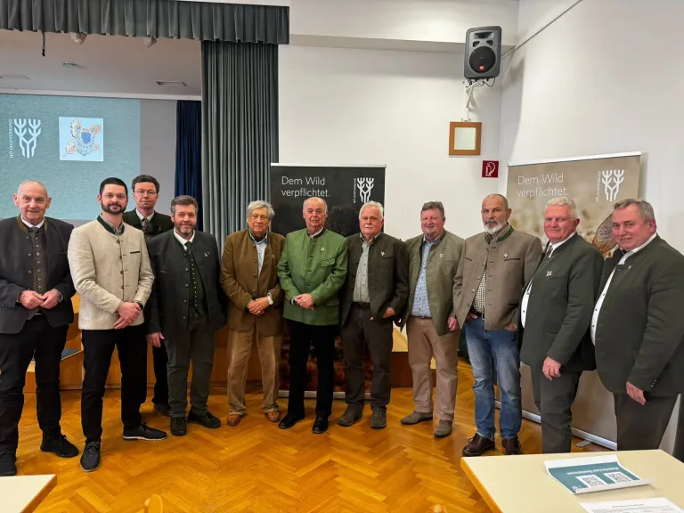 Eine Gruppe von Männern in formeller Kleidung steht in einem Raum mit Holzboden und lächelt für ein Foto. Hinter ihnen befindet sich ein Banner mit dem Text 'Dem Wild verpflichtet'.