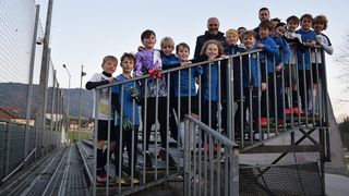 A group of young soccer players in blue uniforms stand on the bleachers, leaning on the railing. A man stands behind them, and some people are standing on the stairs.