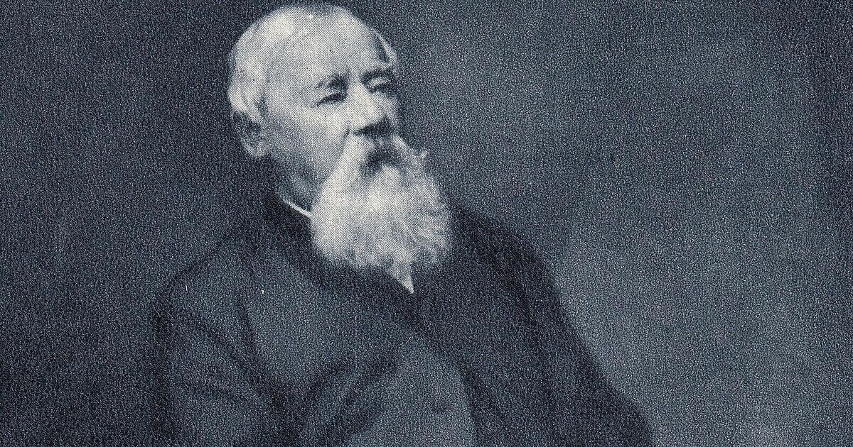 Black and white portrait of an elderly man with a white beard and mustache. He wears a dark jacket and vest.