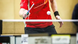 A man wearing a red shirt with 'Sotub' and 'Donau Whirlpools' texts, glasses, and a wristband is playing table tennis, hitting a white ball with a racket.
