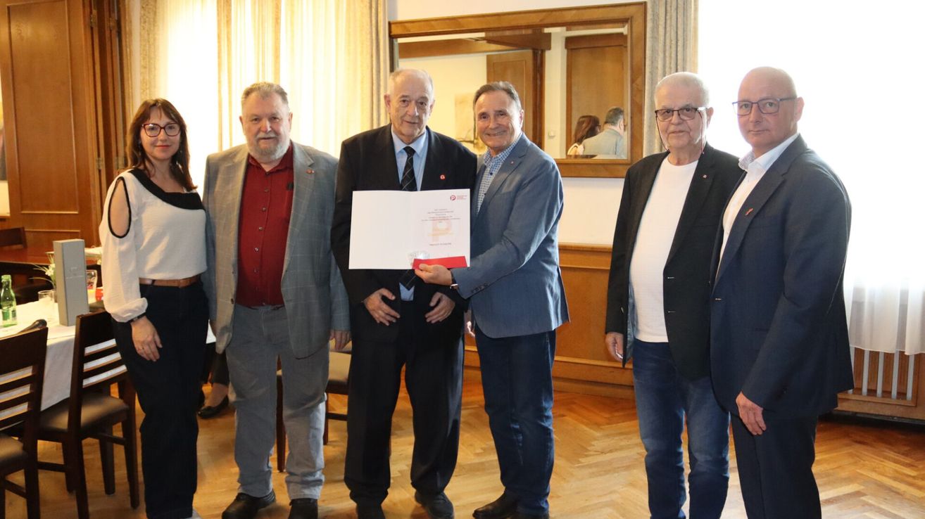 Five men stand in a room with wooden floors and a wooden wall. One man holds a paper with a red logo.