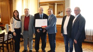 Five men stand in a room with wooden floors and a wooden wall. One man holds a paper with a red logo.