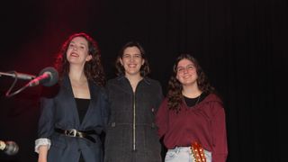 Three women smiling and standing together, one holding a guitar, in front of a black backdrop.