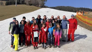 A group of people, adults and children, dressed in red and blue ski suits, pose for a photo in the snow. Trees and a grassy area are in the background.