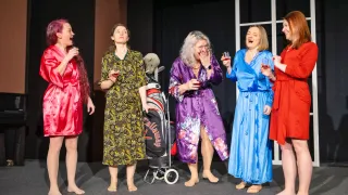 Five women on a stage, all in robes, laugh and hold glasses. The woman in the middle wears purple, with a golf bag behind her.