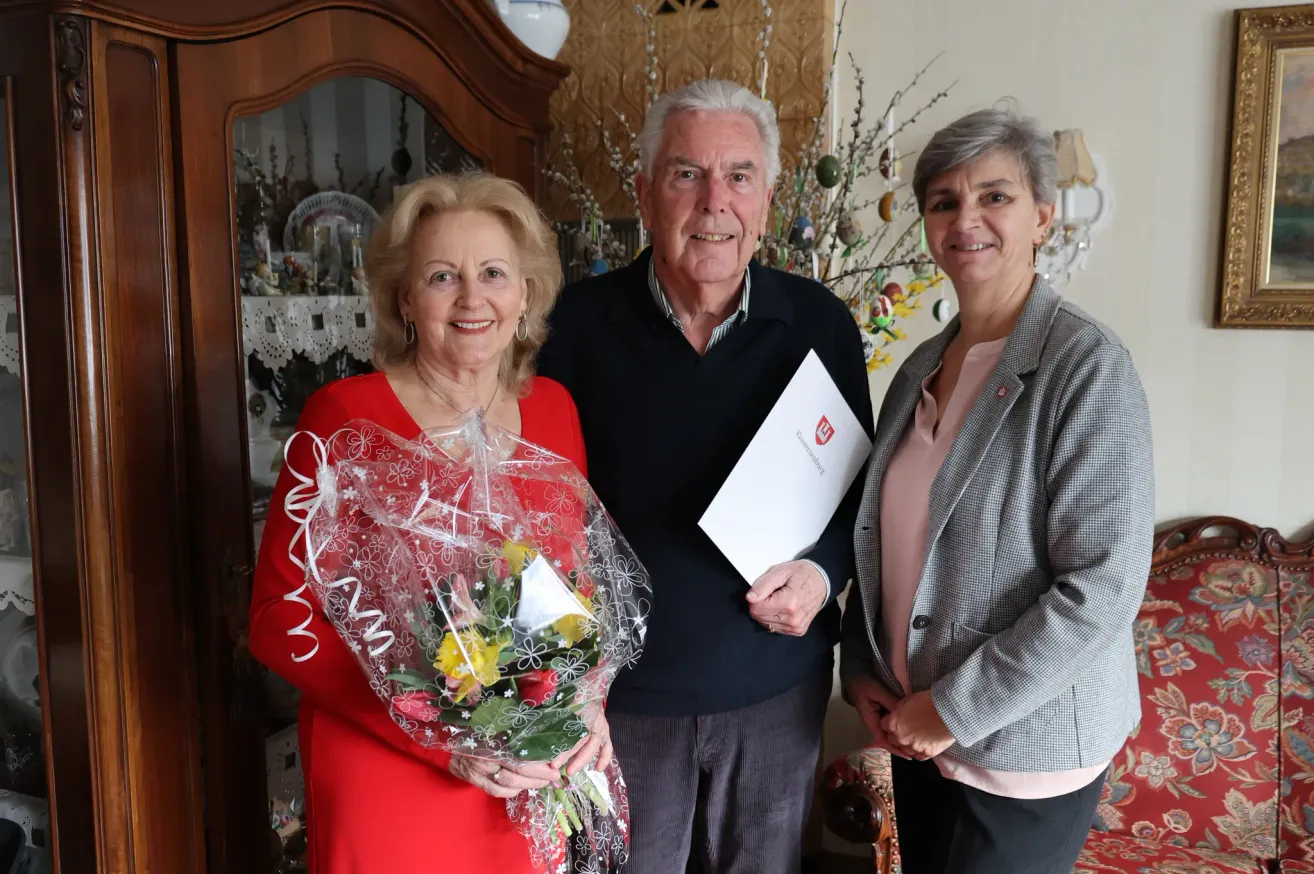Three people stand together in a room, smiling. A woman in red holds a bouquet of flowers, and a man in black holds a certificate. Behind them is a cabinet with decorative items.