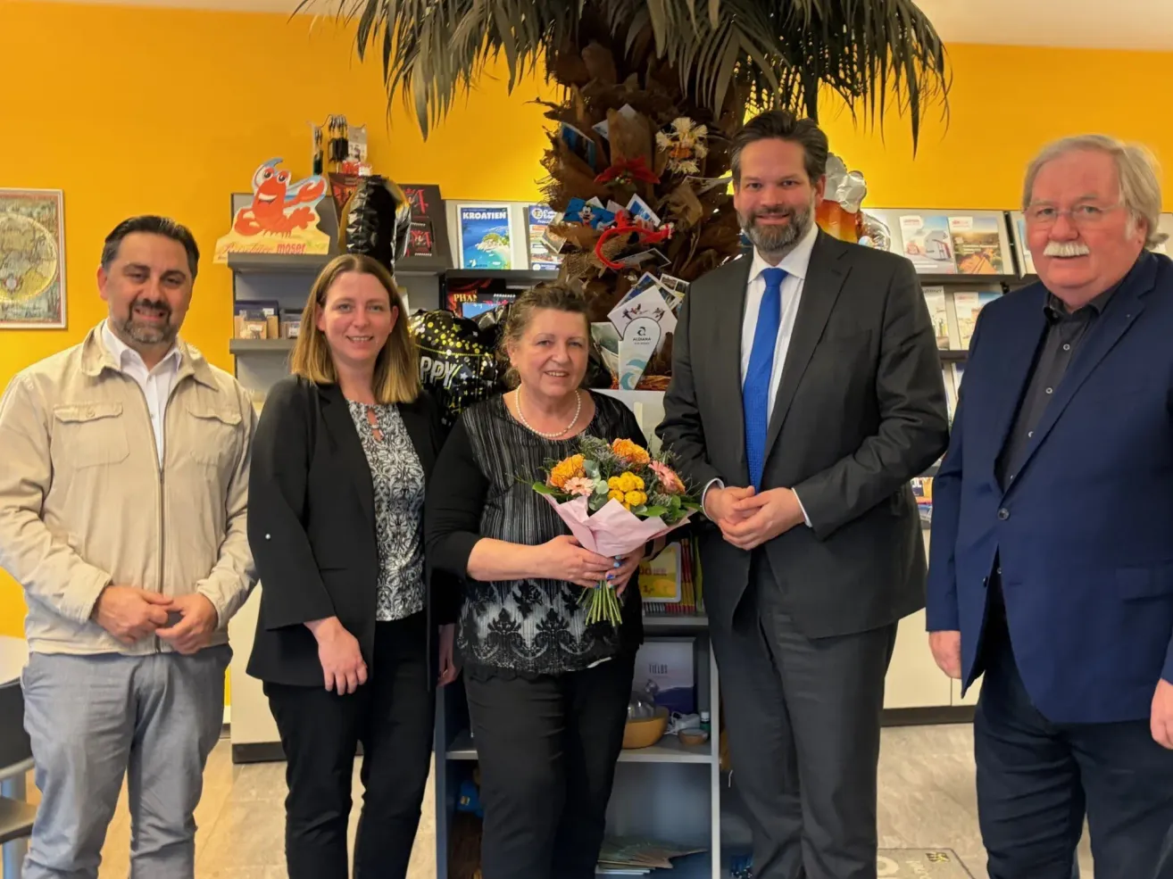 Five people stand together in a room with a yellow wall, a large palm tree decoration, and a shelf with books and items. The woman in the center holds a bouquet of flowers.