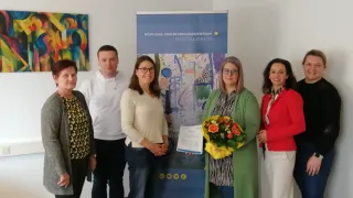 Eine Gruppe von Menschen steht lächelnd vor einem Banner. Eine Frau in der Mitte hält einen Blumenstrauß und ein Zertifikat. Der Raum hat weiße Wände und einen Holzboden.