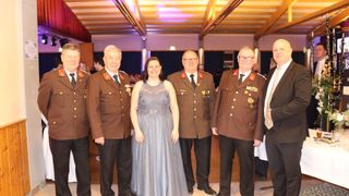 Five people in uniform stand together, smiling. Three men and one woman. The men wear medals and ties. The woman wears a dress. They are likely at a formal event.