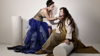 A woman in a gold and blue dress is leaning over a man sitting on a white cylinder. The man has long hair and a beard.