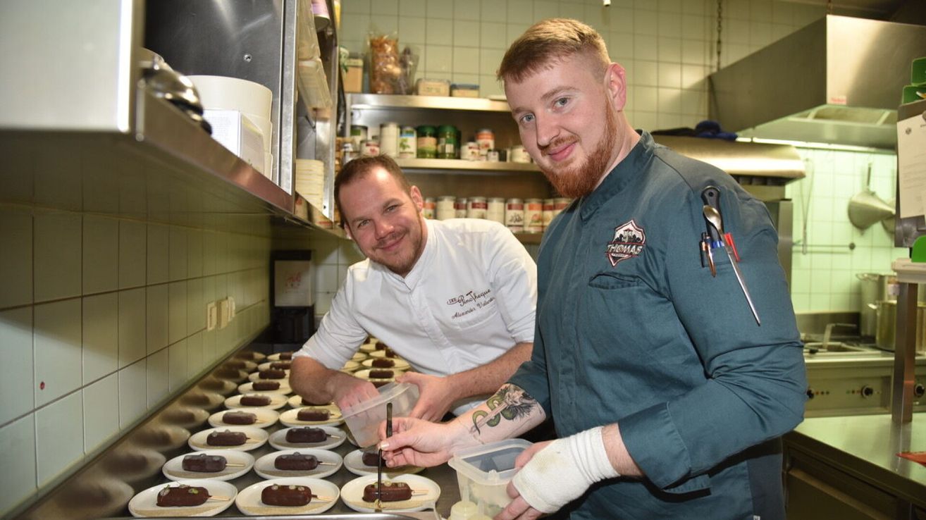 Two chefs in a kitchen, one in a white uniform and the other in a green shirt with a logo, are preparing chocolate popsicles. They have a tray of dessert plates in front of them.