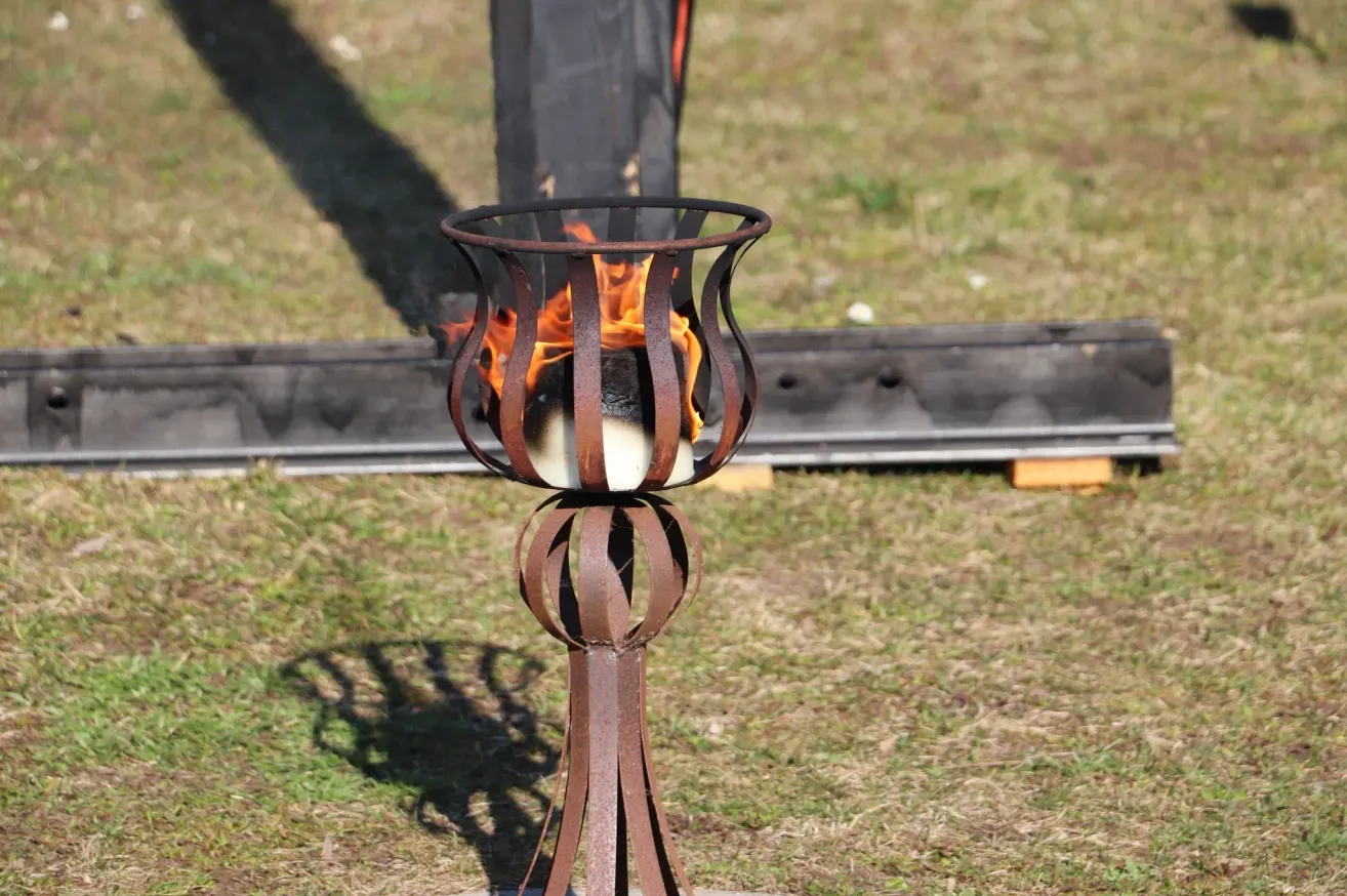 A rustic metal torch stands on a grassy field, with a burning fire inside, casting shadows on the ground.