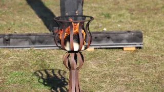 A rustic metal torch stands on a grassy field, with a burning fire inside, casting shadows on the ground.