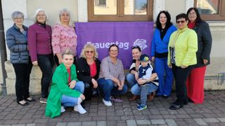 Eine Gruppe von Frauen und einem Kind posiert für ein Foto vor einem lilafarbenen Banner, das Frauentag sagt. Sie tragen verschiedene Outfits und einige haben Brillen. Die Frau in der Mitte hält ein Baby.