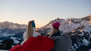 Zwei Frauen in Winterkleidung sitzen auf Bohnensäcken und beobachten einen Sonnenuntergang über verschneiten Bergen. Eine hält ein Weinglas, die andere ein Bierglas.