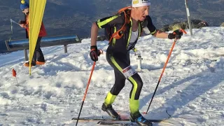 Ein Mann rennt im Rennen bergauf auf Skiern, trägt eine Stirnband, Handschuhe und Skischuhe. Er hält Skistöcke und trägt ein nummeriertes Trikot. Dahinter hält eine Person eine gelbe Flagge.