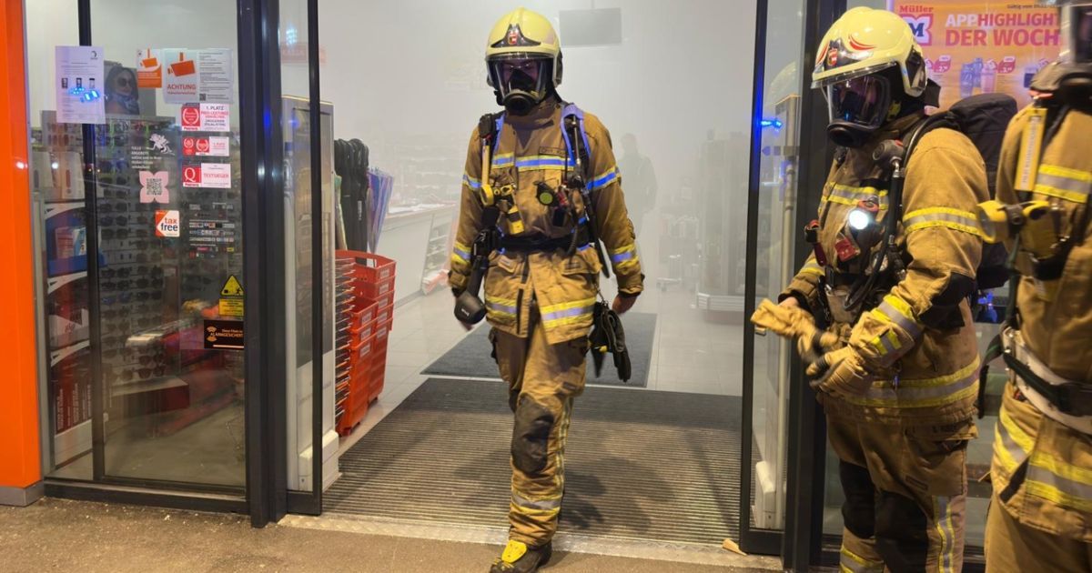 Two firefighters in full gear stand in the entrance of a building. One is holding a tool, and the other looks to the side. Behind them, a blurry scene shows shelves with red storage boxes.