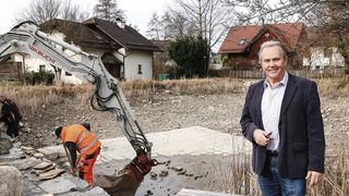 A man in a suit stands near a construction site. An excavator is working on the site, and a worker is standing in front of it. Behind the excavator, there are houses.
