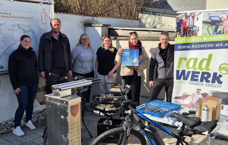 Fünf Personen stehen im Freien mit Fahrrädern. Eine Person hält ein Buch. In der Nähe sind Werkzeuge auf einem Tisch ausgelegt. Ein Banner ist rechts sichtbar.