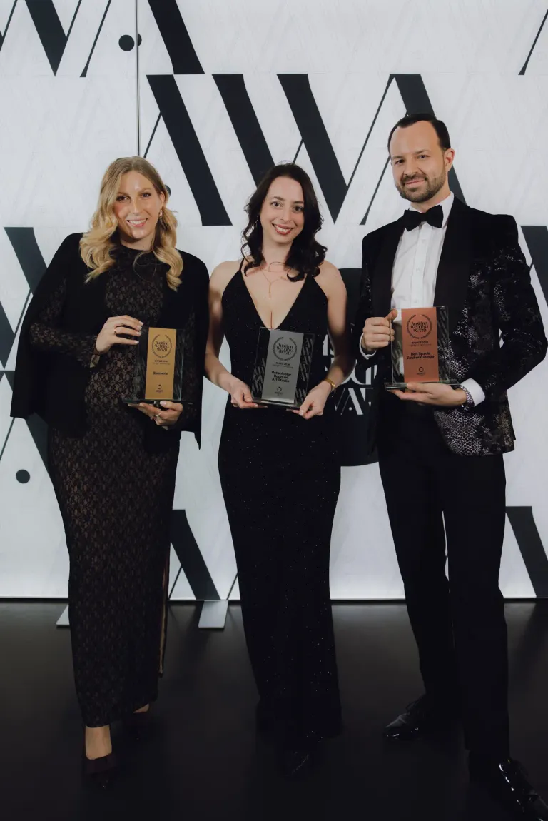 Drei Personen, zwei Frauen und ein Mann, stehen zusammen und halten Auszeichnungen. Die Frauen tragen schwarze Kleider, eines mit Spitze, während der Mann einen Anzug trägt. Sie alle lächeln für ein Foto.