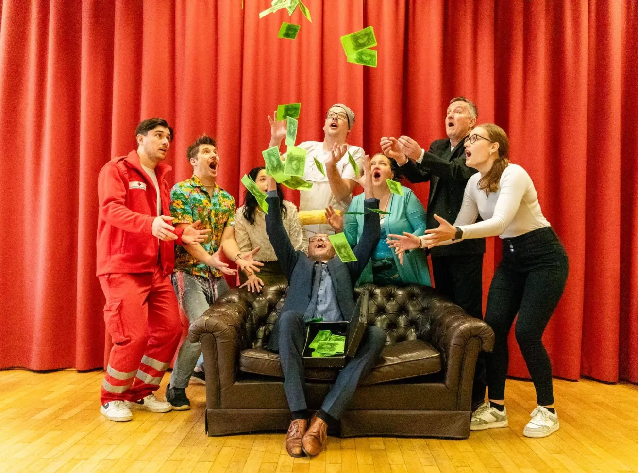 Eine Gruppe von Menschen auf der Bühne, einige werfen Geld, während andere jubeln. Eine Person sitzt auf einem Sofa mit einer Aktentasche voller Geld.