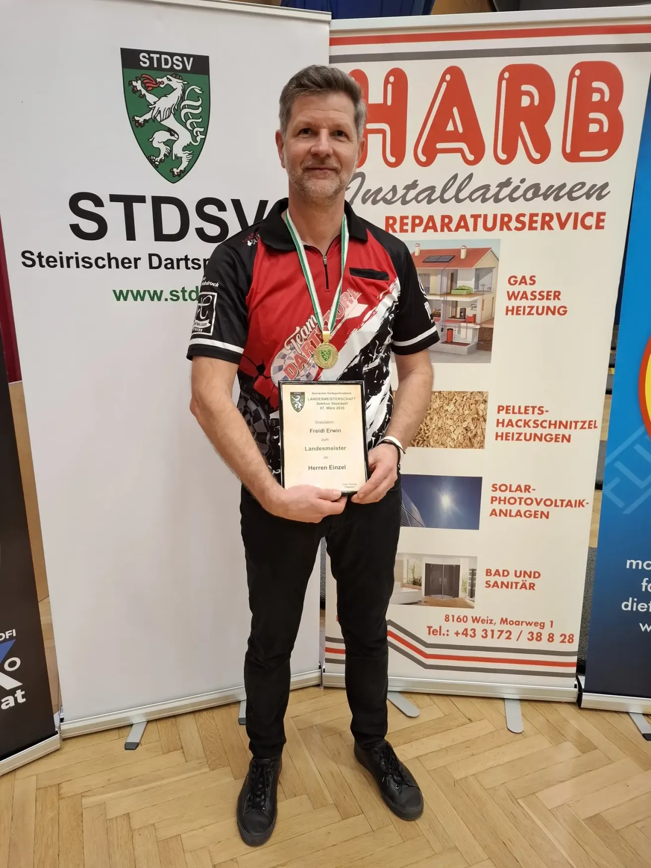 A man stands in front of a banner holding a medal and an award. He wears a red and black shirt, black pants, and shoes. The banner includes text related to Steiermark Darts and installation services.
