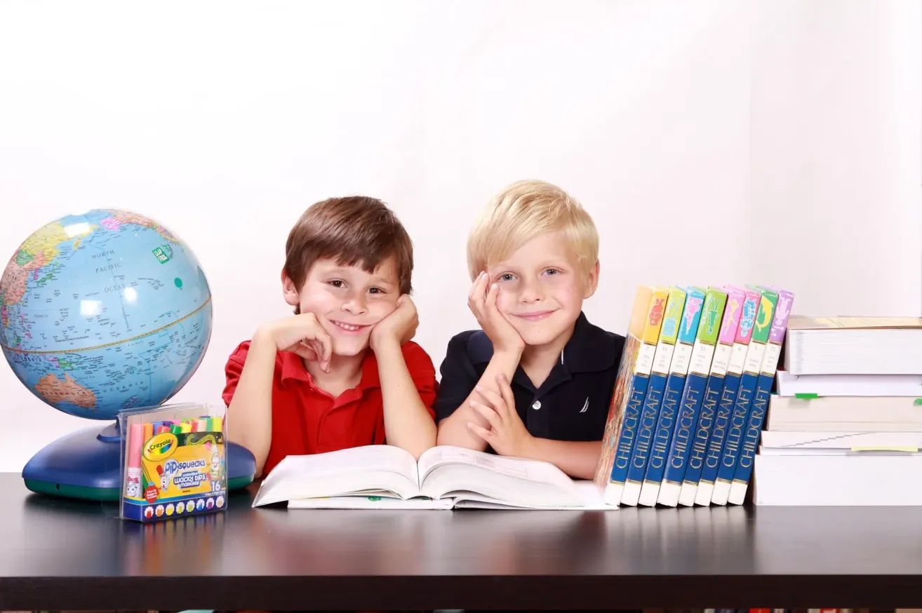 Two boys sitting at a desk with books and crayons, one wearing a red shirt, the other a black polo, smiling at the camera.