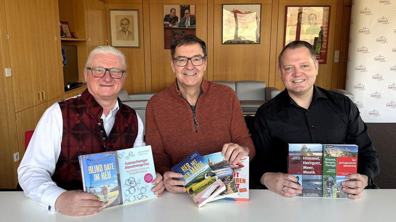 Three men sit at a desk smiling and holding books, one holding a book titled Blind Date im Heu. Behind them, a wall with framed portraits.