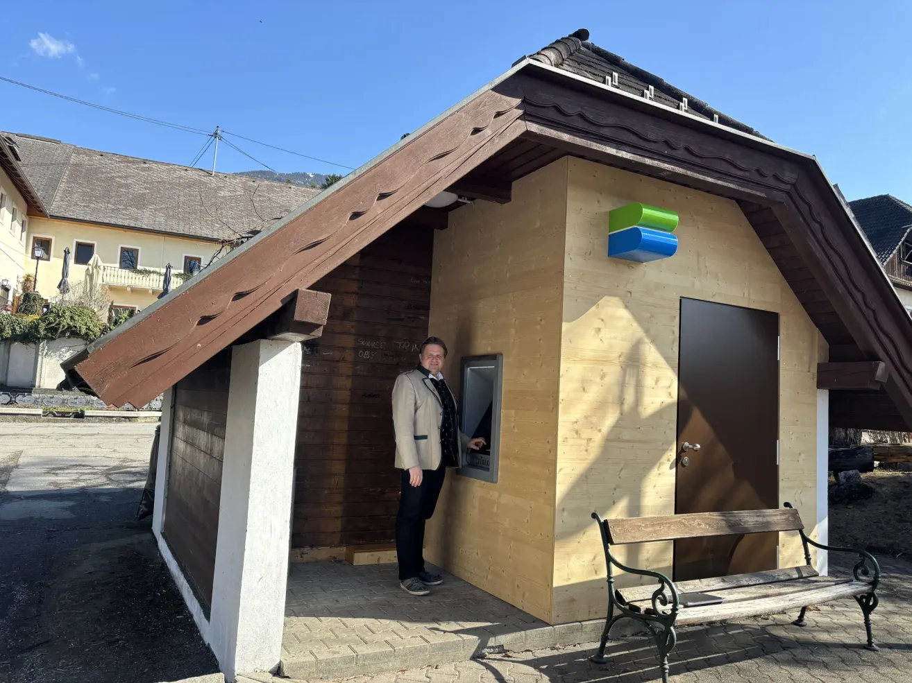 Ein Mann in formeller Kleidung steht an einem Geldautomaten an einem sonnigen Tag, mit einer kleinen Holzbank rechts und einem Holzhaus im Hintergrund.