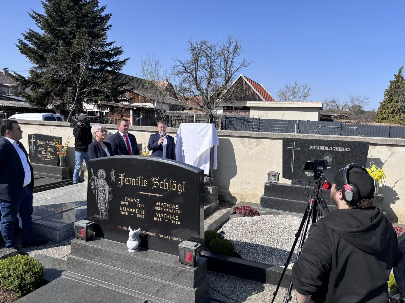 Eine Friedhofsszene mit einer Gruppe von Menschen, die in der Nähe eines Gedenksteins stehen, einer Kamera auf einem Stativ und Bäumen und Gebäuden im Hintergrund.