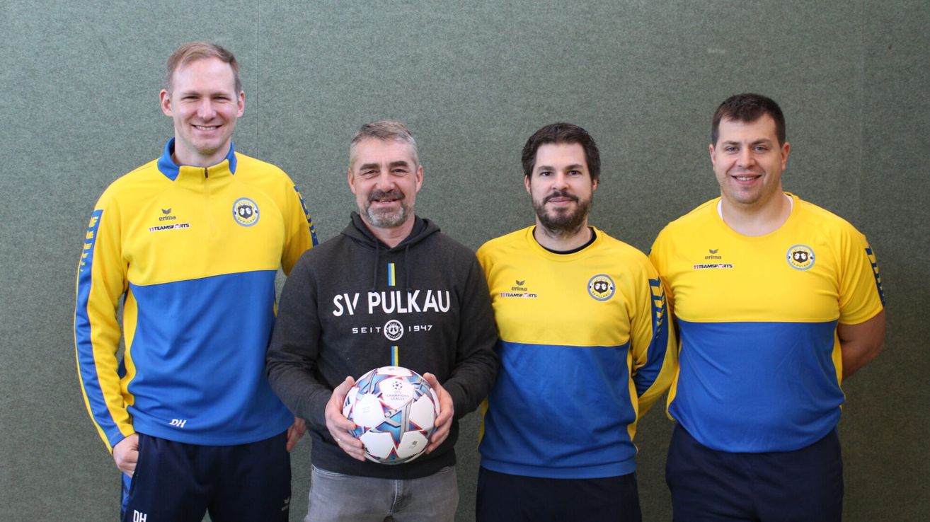 Vier Männer stehen zusammen und lächeln für ein Foto. Der Mann in der Mitte hält einen Fußball. Sie tragen alle Trikots in Gelb und Blau.