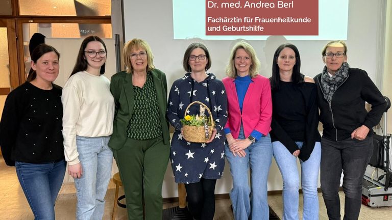 Fünf Frauen stehen vor einer Leinwand mit der Aufschrift 'Dr. med. Andrea Berl, Gynäkologin für Frauengesundheit und Geburtshilfe.' Eine Frau hält einen Korb mit Blumen.