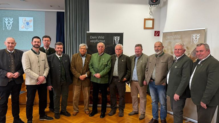 Eine Gruppe von Männern in traditioneller Kleidung steht in einem Raum mit Holzböden. Sie lächeln und scheinen für ein Foto zu posieren. Hinter ihnen befindet sich ein Banner mit Text in deutscher Sprache.