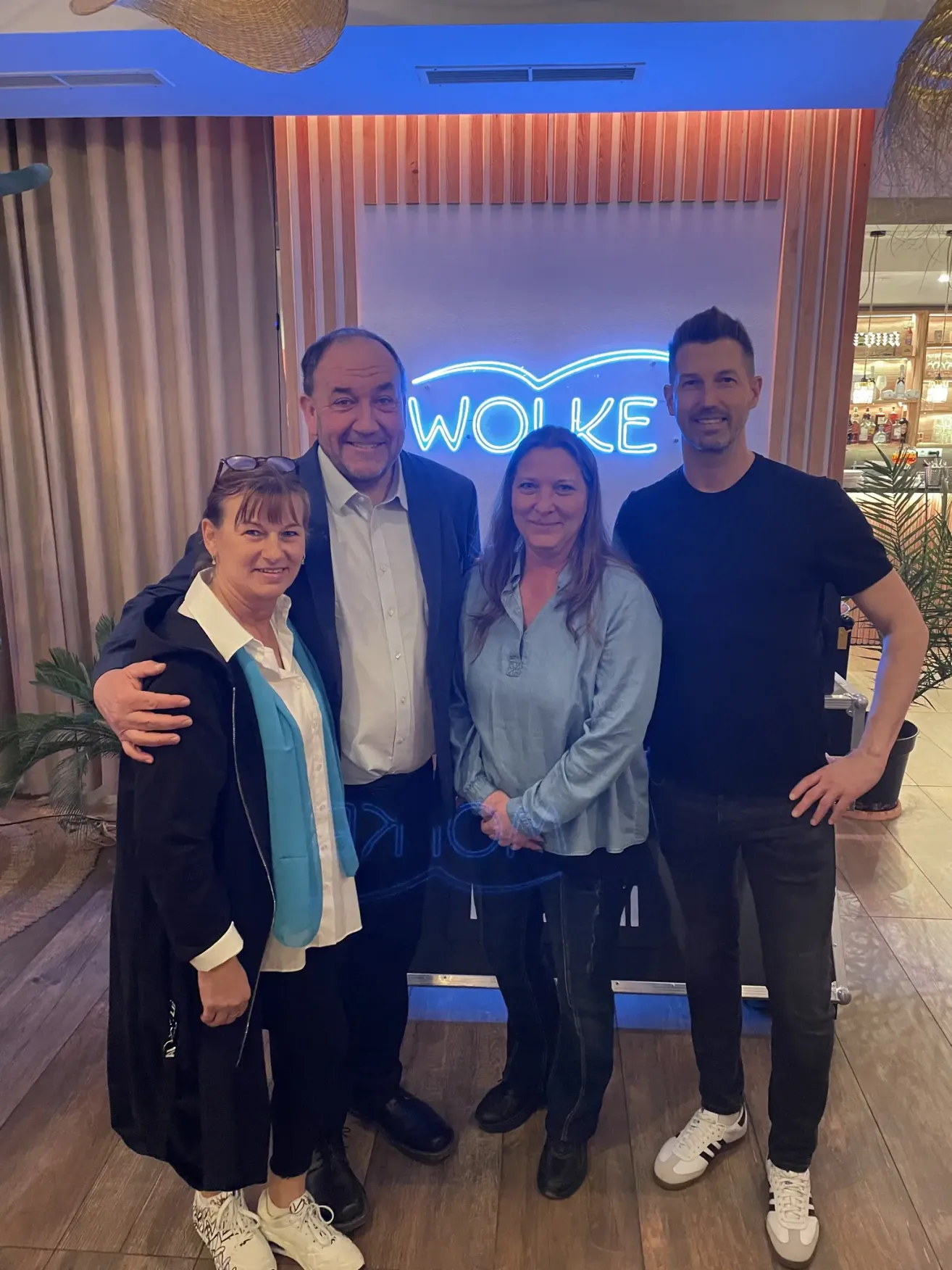 Four people pose in front of a neon sign that reads 'WOLKE'. Two women and two men, smiling, with one man in a suit and the other in a black t-shirt. Behind them, a bar setting is visible.