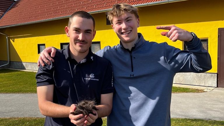 Zwei Männer posieren für ein Foto vor einem gelben Gebäude. Ein Mann hält einen Vogel, während der andere deutet und lächelt.
