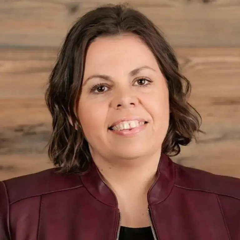 Eine Frau mit kurzen, lockigen braunen Haaren lächelt. Sie trägt eine bordeauxfarbene Lederjacke. Der Hintergrund ist eine Holzwand.
