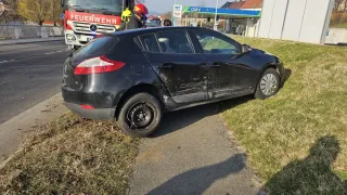 Ein schwarzer Renault Clio Kombi ist am Straßenrand beschädigt. Zwei Personen stehen in der Nähe eines Feuerwehrwagens. Ein Tankstellengebäude ist im Hintergrund zu sehen.