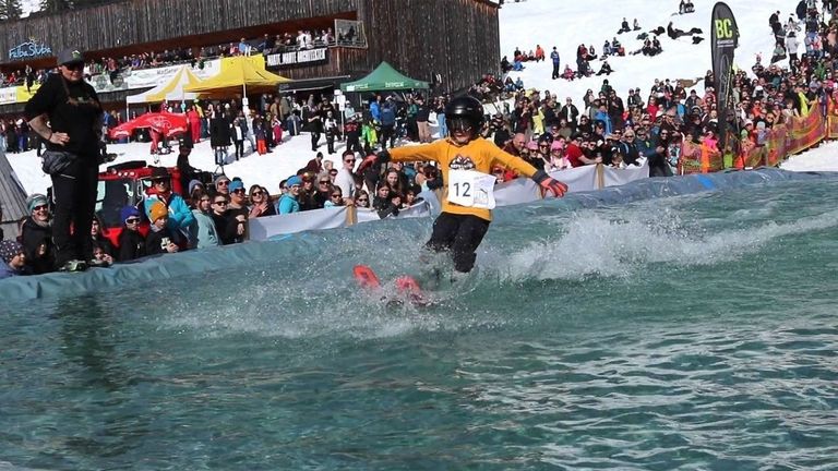 Ein Skifahrer in einem gelben Shirt und Helm mit der Nummer 12 springt in einen Pool, während Zuschauer an den schneebedeckten Seitenlinien zusehen.