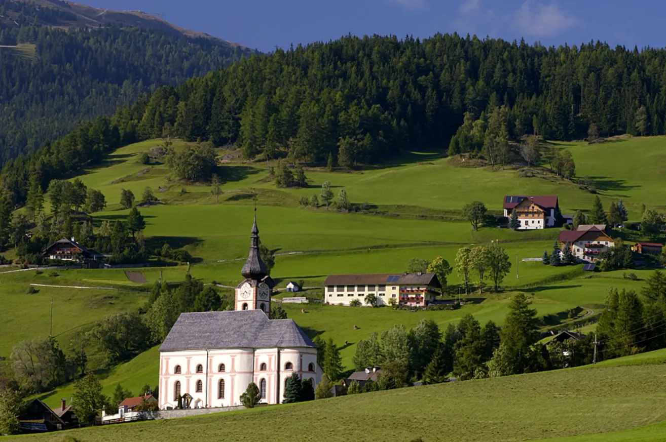 Ein malerisches Bild einer Kirche mit einem Turm, umgeben von saftig grünen Wiesen und dichten Wäldern unter einem blauen Himmel.