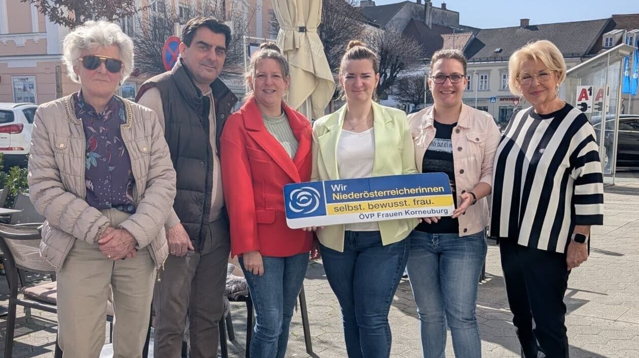 A group of four people, three women and one man, are standing close together, smiling for a photo. They hold a blue banner with German text. The background shows a city street with trees and buildings.