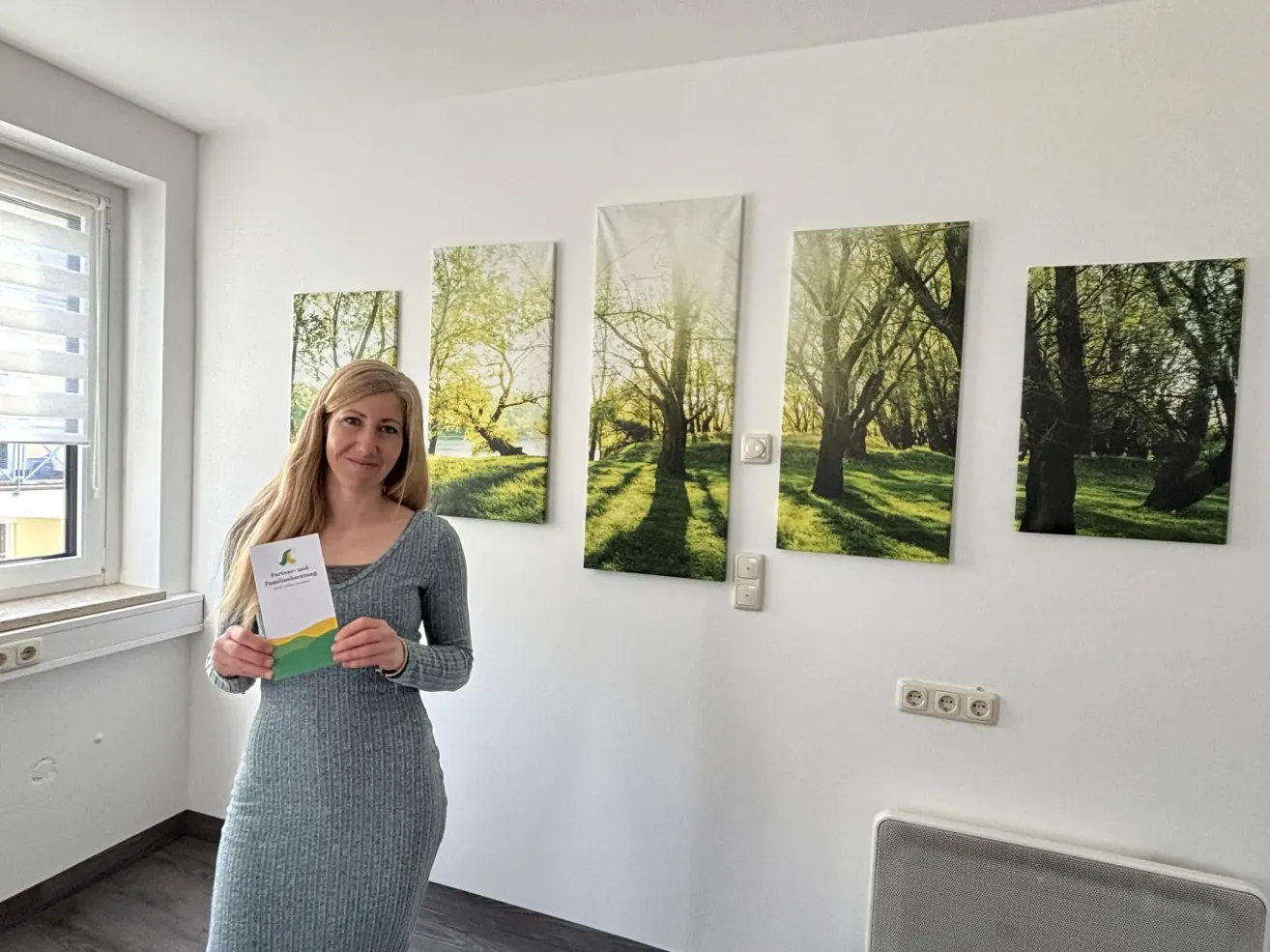 Eine Frau in einem grauen Kleid hält ein Büchlein vor einer Wand mit vier Landschaftsgemälden. Sie lächelt und steht neben einem Fenster.