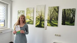 Eine Frau in einem grauen Kleid hält ein Büchlein vor einer Wand mit vier Landschaftsgemälden. Sie lächelt und steht neben einem Fenster.