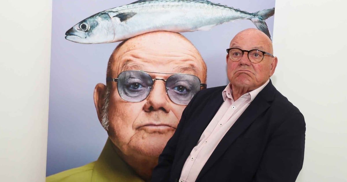 An older man stands before a poster of himself wearing sunglasses with fish eyes, a fish on his head, and a serious expression. He wears a black suit and glasses.