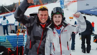 Zwei Skifahrer feiern auf einem verschneiten Hang und tragen weiße Jacken mit rotem Saum. Der Mann hat eine Tarnjacke und eine Sonnenbrille auf dem Kopf, während die Frau eine Schutzbrille trägt. Hinter ihnen ist ein schneebedeckter Berg und eine Skiliftanlage zu sehen.
