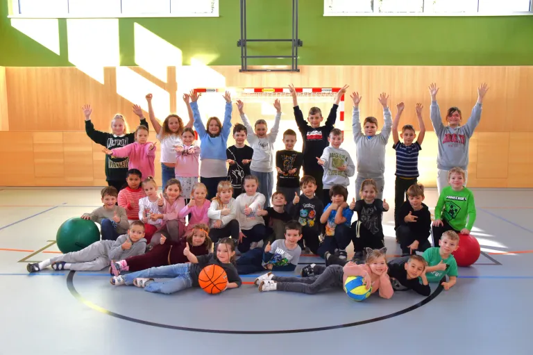 Eine Gruppe von Kindern und Erwachsenen posiert für ein Foto in einer Turnhalle, einige halten Basketballs, und alle zeigen Daumen hoch.
