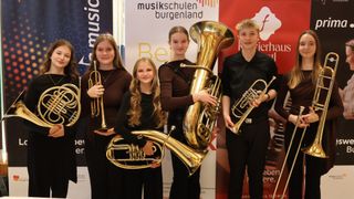 Vier junge Musiker stehen vor einem Banner der Musikschule. Sie halten verschiedene Blechblasinstrumente, darunter eine Trompete, ein Tuba und ein Posaune. Das Banner steht für 'musikschulen burgenland'.