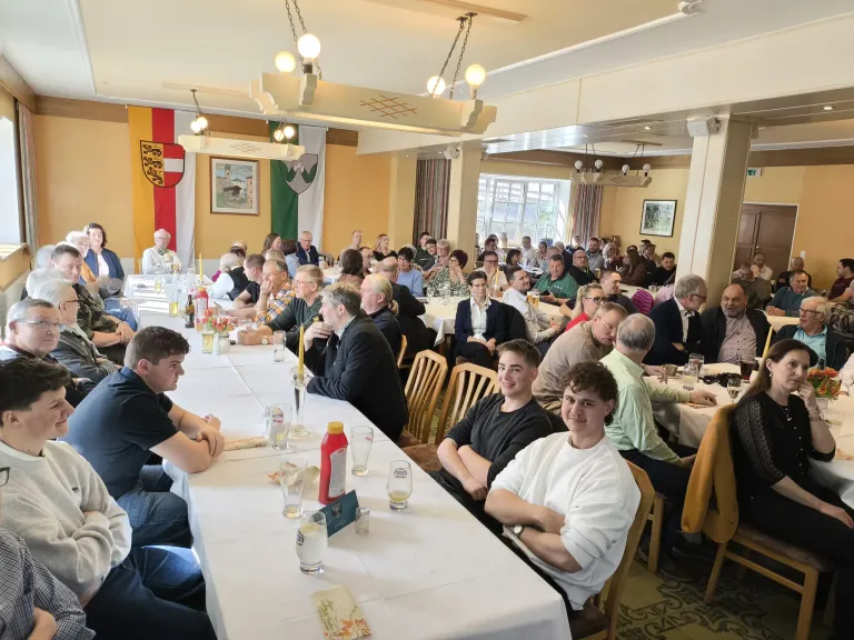 Eine große Gruppe von Menschen ist in einem Bankettsaal versammelt, an langen Tischen sitzend. Der Raum ist mit Flaggen und Bildern an den Wänden dekoriert. Einige unterhalten sich, während andere umherschauen. Verschiedene Gegenstände wie Flaschen, Gläser und Blumen stehen auf den Tischen.